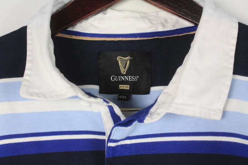 Puede incluir: Una camiseta de rugby a rayas azul marino, azul claro y blanco con cuello blanco. La camiseta presenta el logotipo de Guinness en una etiqueta negra. Talla 4XL.