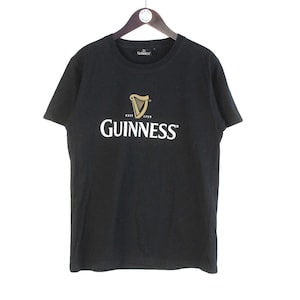以下が含まれることがあります： 黒いTシャツ。白と金色のギネスのロゴ入り。ロゴはハープと太字の「Guinness」の文字。木製のハンガーにかかっています。