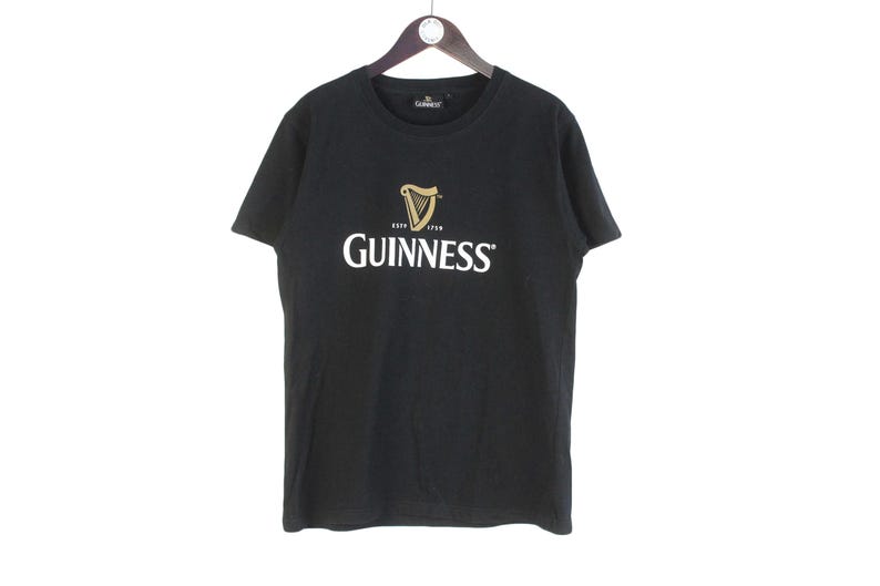 以下が含まれることがあります： 黒いTシャツ。白と金色のギネスのロゴ入り。ロゴはハープと太字の「Guinness」の文字。木製のハンガーにかかっています。