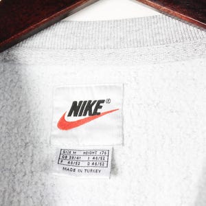 Puede incluir: Sudadera gris con un logotipo de Nike blanco y una etiqueta. La etiqueta dice "Talla M", "Altura 178", "GB 38/41", "I 46/52", "F 48/52", "D 48/52" y "Hecho en Turqu&iacute;a".