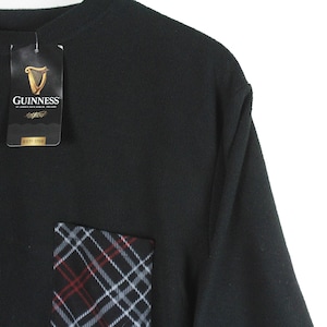 Op de afbeelding: Zwart T-shirt met een geruit zakje en een Guinness-label. De zak heeft een patroon van rode, witte en zwarte lijnen. Het label toont het Guinness-logo en merkinformatie.