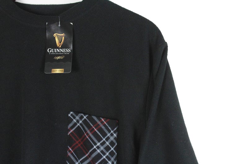 Op de afbeelding: Zwart T-shirt met een geruit zakje en een Guinness-label. De zak heeft een patroon van rode, witte en zwarte lijnen. Het label toont het Guinness-logo en merkinformatie.