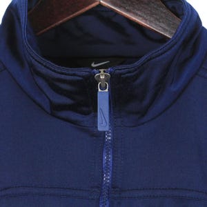 May include: Close-up of a navy blue athletic jacket with a high collar and a full zipper. The jacket features the Nike logo on the chest and zipper pull. The jacket is hanging on a wooden hanger.