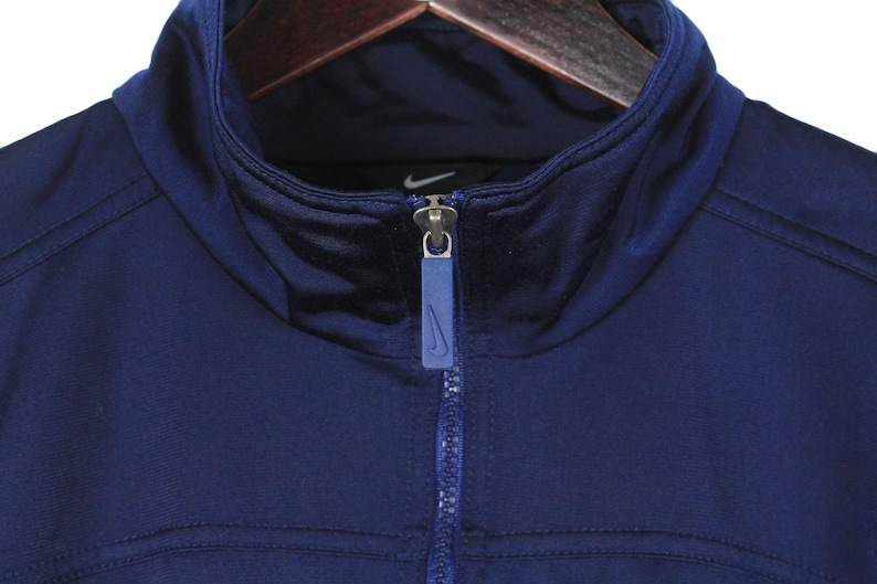 May include: Close-up of a navy blue athletic jacket with a high collar and a full zipper. The jacket features the Nike logo on the chest and zipper pull. The jacket is hanging on a wooden hanger.