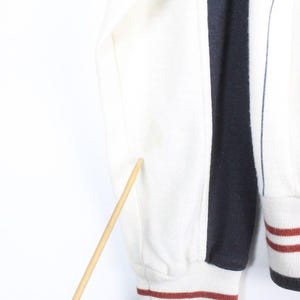 May include: Close-up of a section of a striped sweatshirt. The sweatshirt features alternating vertical stripes of white and navy blue. The sleeve cuff and bottom hem have a white base with red and navy stripes. The sleeve is white.