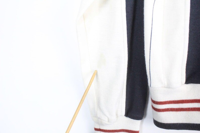 May include: Close-up of a section of a striped sweatshirt. The sweatshirt features alternating vertical stripes of white and navy blue. The sleeve cuff and bottom hem have a white base with red and navy stripes. The sleeve is white.