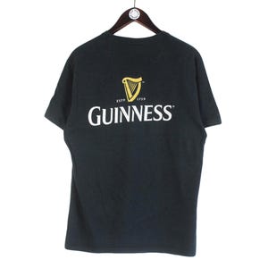 Pu&ograve; includere: T-shirt nera con il logo Guinness bianco e oro. Il logo presenta un'arpa e la scritta "Guinness". La maglietta &egrave; appesa a una gruccia di legno.