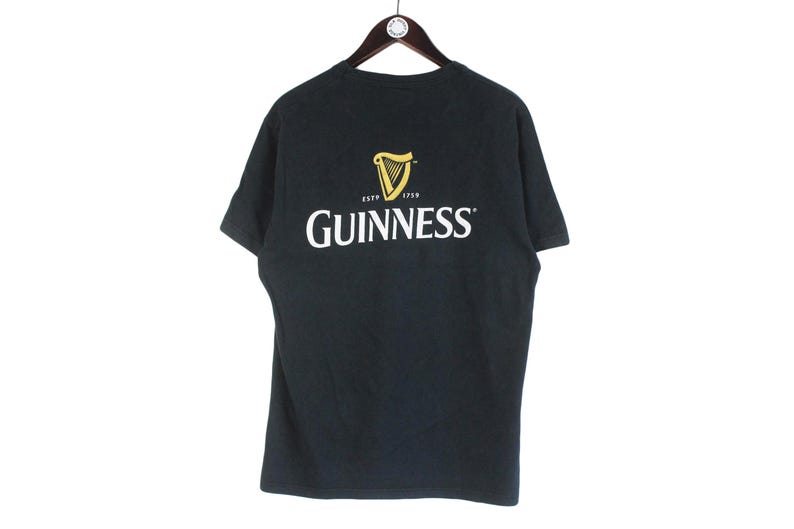 Pu&ograve; includere: T-shirt nera con il logo Guinness bianco e oro. Il logo presenta un'arpa e la scritta "Guinness". La maglietta &egrave; appesa a una gruccia di legno.