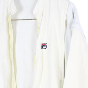 May include: A white zip-up jacket featuring a high collar and long sleeves. The garment displays a small embroidered Fila logo on the left chest area, rendered in navy blue, red, and white thread. The jacket has a central zipper closure.