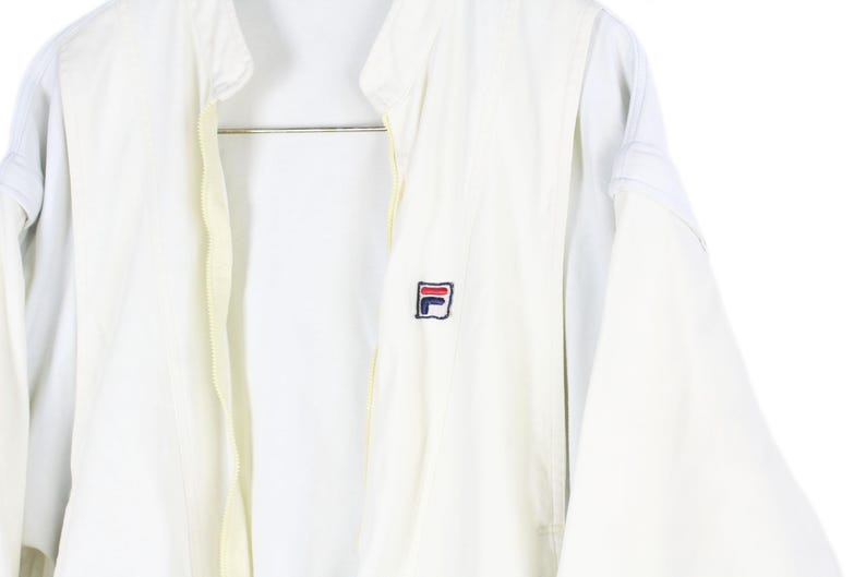 May include: A white zip-up jacket featuring a high collar and long sleeves. The garment displays a small embroidered Fila logo on the left chest area, rendered in navy blue, red, and white thread. The jacket has a central zipper closure.