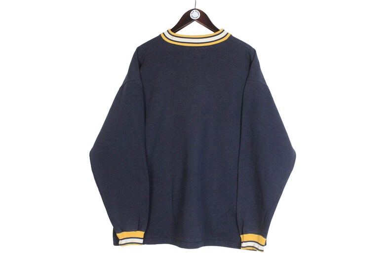 K&ouml;nnte beinhalten: Marineblaues Langarm-Sweatshirt mit gelb-wei&szlig;en Zierleisten an Kragen und B&uuml;ndchen. Das Sweatshirt h&auml;ngt an einem Holzb&uuml;gel vor wei&szlig;em Hintergrund. Das Kleidungsst&uuml;ck hat einen klassischen, l&auml;ssigen Stil.