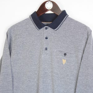 May include: A gray long-sleeve polo shirt with a navy blue collar and cuffs. The shirt has a three-button placket, a chest pocket, and a small embroidered emblem. The collar has a white trim.