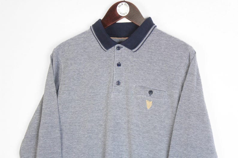 May include: A gray long-sleeve polo shirt with a navy blue collar and cuffs. The shirt has a three-button placket, a chest pocket, and a small embroidered emblem. The collar has a white trim.