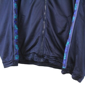 Puede incluir: Una chaqueta azul marino con cremallera y ribete a rayas azul y verde. El ribete tiene las palabras "Puma" y "Puma Sport" repetidas a lo largo del ribete.