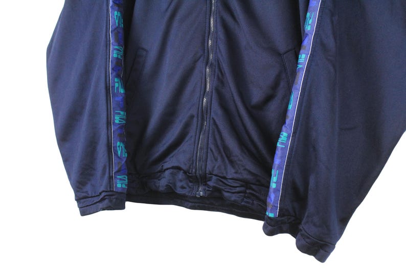 Puede incluir: Una chaqueta azul marino con cremallera y ribete a rayas azul y verde. El ribete tiene las palabras "Puma" y "Puma Sport" repetidas a lo largo del ribete.