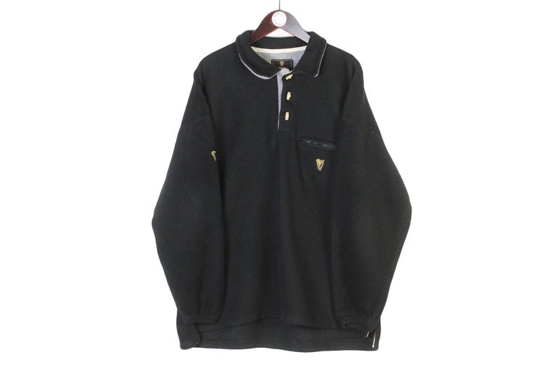 May include: Black long-sleeved pullover shirt with a collared neckline and a three-button placket. Features a small pocket on the chest and a gold-colored emblem. The shirt has a white trim on the collar and cuffs. The Guinness logo is visible.