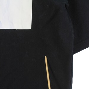 May include: Black t-shirt with a white rectangular design on the upper left. The design appears to be a partial image or graphic. The shirt's fabric is visible, and the sleeves are folded. A wooden stick is in the lower center.