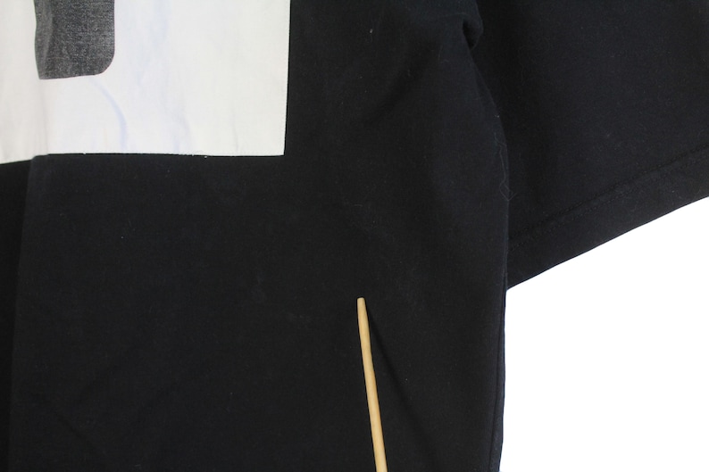 May include: Black t-shirt with a white rectangular design on the upper left. The design appears to be a partial image or graphic. The shirt's fabric is visible, and the sleeves are folded. A wooden stick is in the lower center.