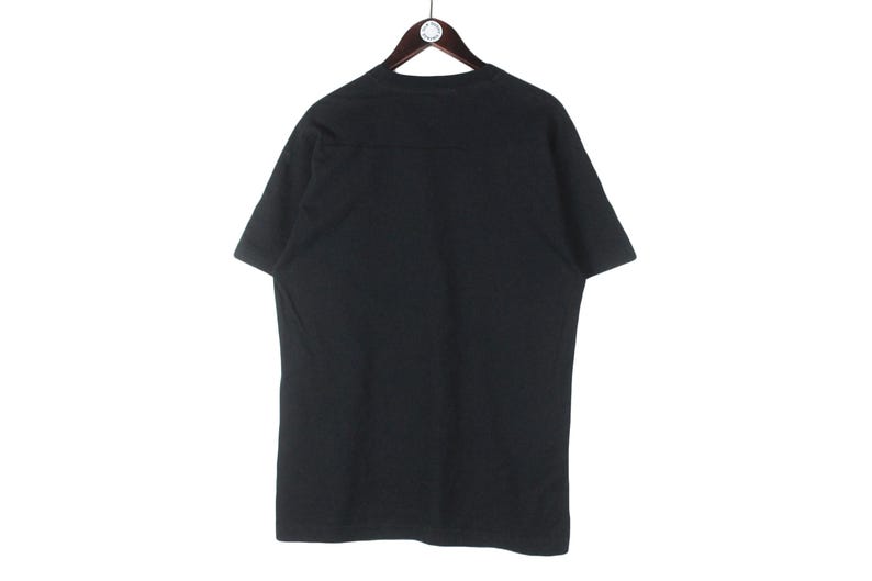Puede incluir: Una camiseta negra lisa colgada de una percha de madera. La camiseta de manga corta est&aacute; hecha de un material suave y tiene un dise&ntilde;o sencillo. La camiseta se muestra sobre un fondo blanco.