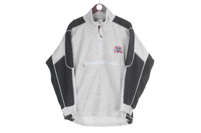 Pu&ograve; includere: Un pullover in pile grigio con pannelli neri sulle spalle e sulle maniche. Presenta una chiusura con zip a un quarto, una piccola tasca sul petto e una toppa con il logo "Police Club California" in rosso, bianco e blu.