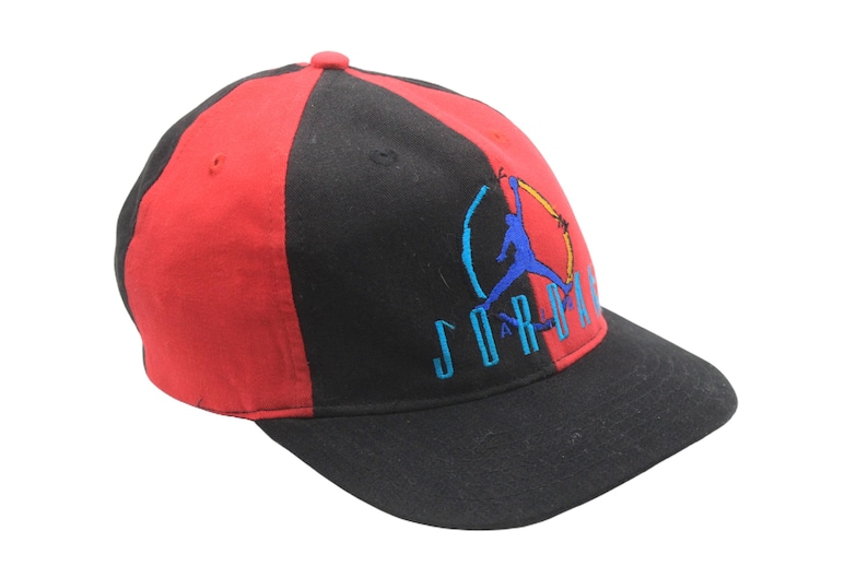 May include: A red and black baseball cap with the Jordan logo embroidered on the front. The cap has a curved brim and a six-panel design. The logo features a silhouette of a basketball player in blue, with the word "JORDAN" in blue.