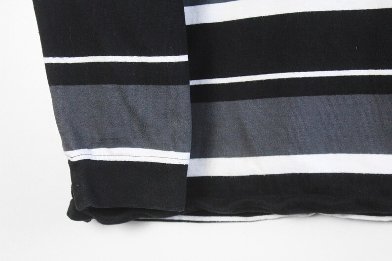 May include: Close-up of a long-sleeved shirt with horizontal stripes. The shirt features alternating stripes of black, white, and gray. The sleeve cuff is solid black. The fabric appears to be a soft knit material.