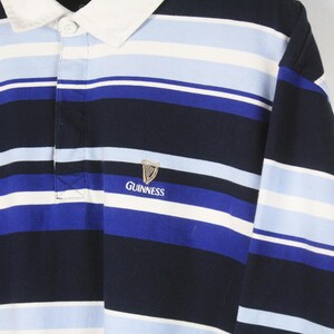 Puede incluir: Una camiseta de rugby de manga larga con rayas horizontales en azul marino, azul claro y blanco. La camiseta tiene un cuello blanco y un peque&ntilde;o logotipo de Guinness bordado en el pecho.