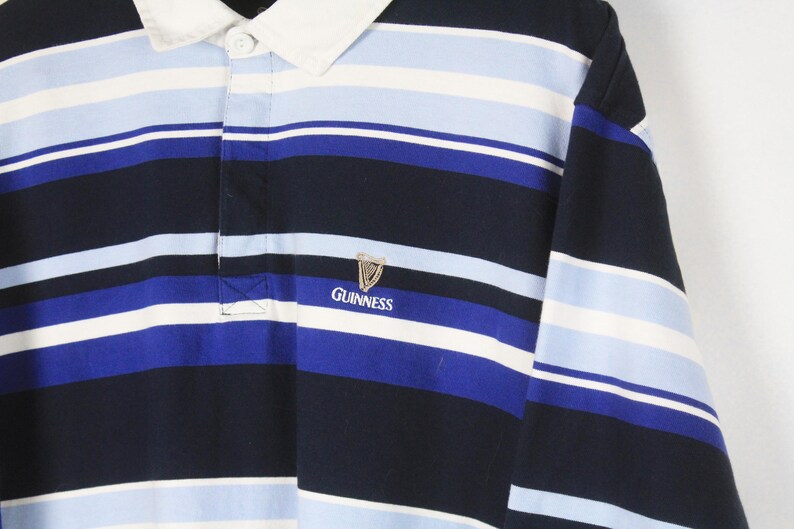 Puede incluir: Una camiseta de rugby de manga larga con rayas horizontales en azul marino, azul claro y blanco. La camiseta tiene un cuello blanco y un peque&ntilde;o logotipo de Guinness bordado en el pecho.