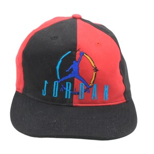 May include: Black and red baseball cap with the word "JORDAN" embroidered in teal. The cap features a blue silhouette of a basketball player. The hat is divided into two color blocks, with black on one side and red on the other.