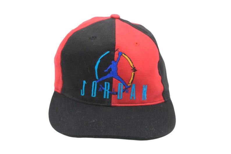 May include: Black and red baseball cap with the word "JORDAN" embroidered in teal. The cap features a blue silhouette of a basketball player. The hat is divided into two color blocks, with black on one side and red on the other.