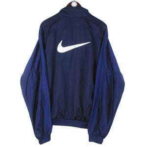 May include: Navy blue Nike track jacket with a white Nike swoosh logo on the back. The jacket has a high collar and long sleeves. The sleeves and upper back are navy blue, while the lower sleeves are a darker shade of blue.