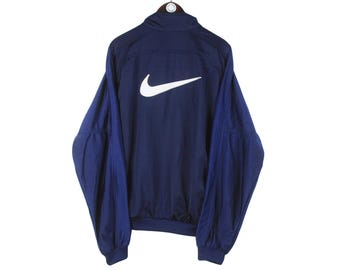 May include: Navy blue Nike track jacket with a white Nike swoosh logo on the back. The jacket has a high collar and long sleeves. The sleeves and upper back are navy blue, while the lower sleeves are a darker shade of blue.