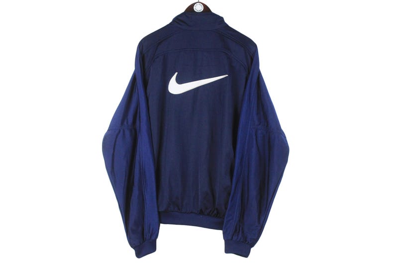May include: Navy blue Nike track jacket with a white Nike swoosh logo on the back. The jacket has a high collar and long sleeves. The sleeves and upper back are navy blue, while the lower sleeves are a darker shade of blue.