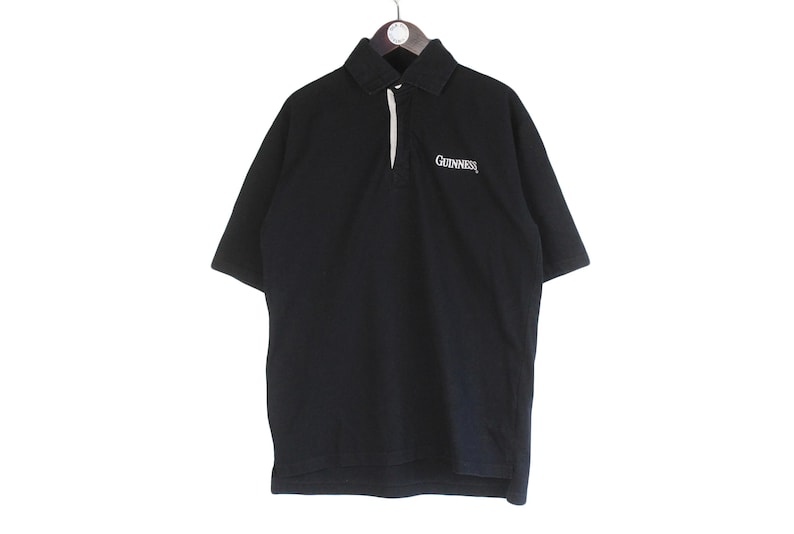 May include: Black polo shirt with short sleeves, a collared neckline, and a white placket. The word "Guinness" is embroidered in white on the chest. The shirt is hanging from a hanger.