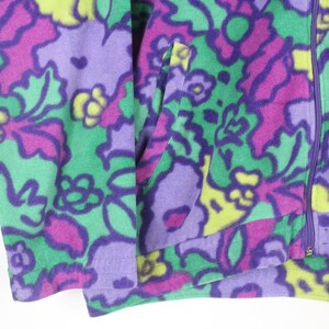 Puede incluir: Una chaqueta de forro polar con un vibrante estampado floral multicolor en tonos morado, verde brillante y amarillo. La prenda incluye un cierre de cremallera morado de longitud completa en el centro y un bolsillo lateral funcional en el panel frontal.