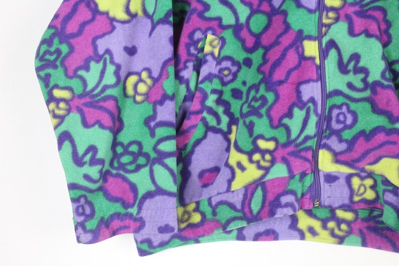 Puede incluir: Una chaqueta de forro polar con un vibrante estampado floral multicolor en tonos morado, verde brillante y amarillo. La prenda incluye un cierre de cremallera morado de longitud completa en el centro y un bolsillo lateral funcional en el panel frontal.