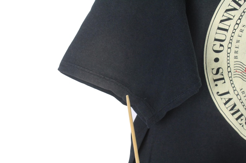 May include: Black t-shirt featuring a partial view of a Guinness logo. The logo is circular with text and a harp design. The shirt's sleeve is visible, showing the fabric's texture.
