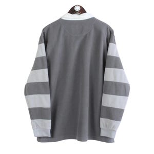Puede incluir: Una camisa gris de manga larga con mangas a rayas blancas y cuello blanco. La camisa est&aacute; colgada en una percha de madera sobre un fondo blanco. La camisa est&aacute; hecha de un material suave.