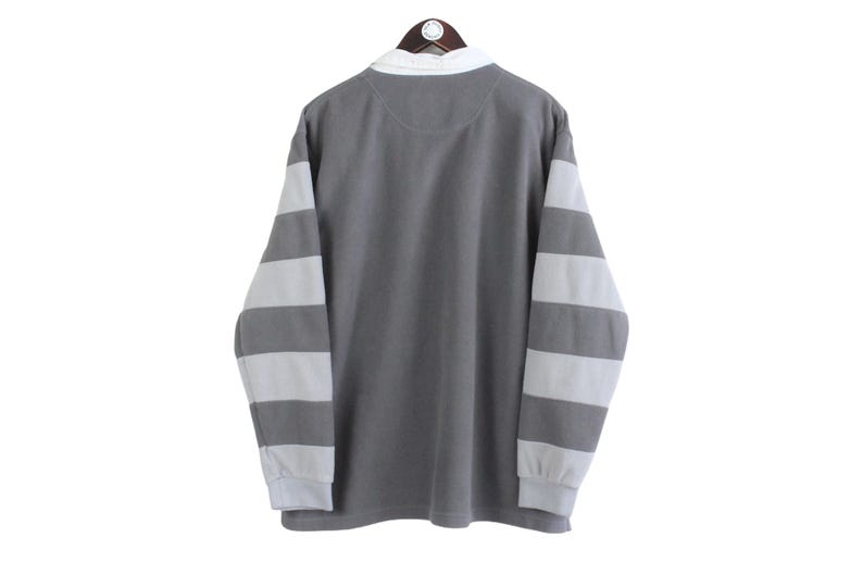 Puede incluir: Una camisa gris de manga larga con mangas a rayas blancas y cuello blanco. La camisa est&aacute; colgada en una percha de madera sobre un fondo blanco. La camisa est&aacute; hecha de un material suave.