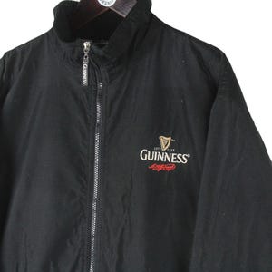 K&ouml;nnte beinhalten: Schwarze Rei&szlig;verschlussjacke mit Fleecefutter und einem gestickten Guinness-Logo auf der Vorderseite. Das Logo ist rot und wei&szlig; mit einer schwarzen Harfe.