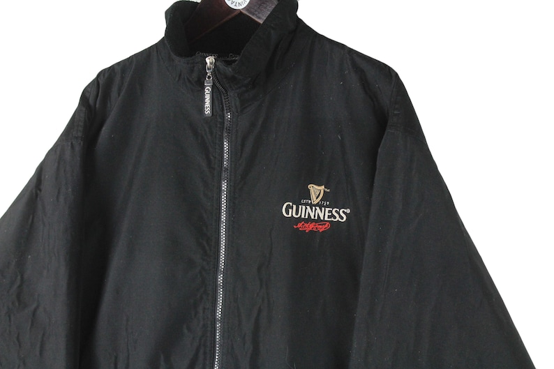 K&ouml;nnte beinhalten: Schwarze Rei&szlig;verschlussjacke mit Fleecefutter und einem gestickten Guinness-Logo auf der Vorderseite. Das Logo ist rot und wei&szlig; mit einer schwarzen Harfe.