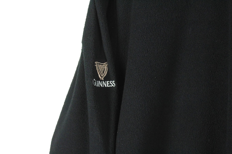 Op de afbeelding: Zwarte fleecejas met het Guinness-logo geborduurd op de mouw. Het logo toont een gouden harp en het woord "Guinness" in een witte, gestileerde letter. De jas heeft een getextureerde uitstraling.