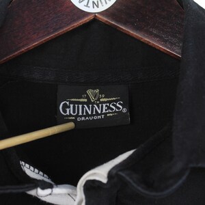 May include: Close-up of a black Guinness Draught shirt collar. The shirt has a white collar and a black label with the Guinness logo and text. The shirt is hanging on a wooden hanger.