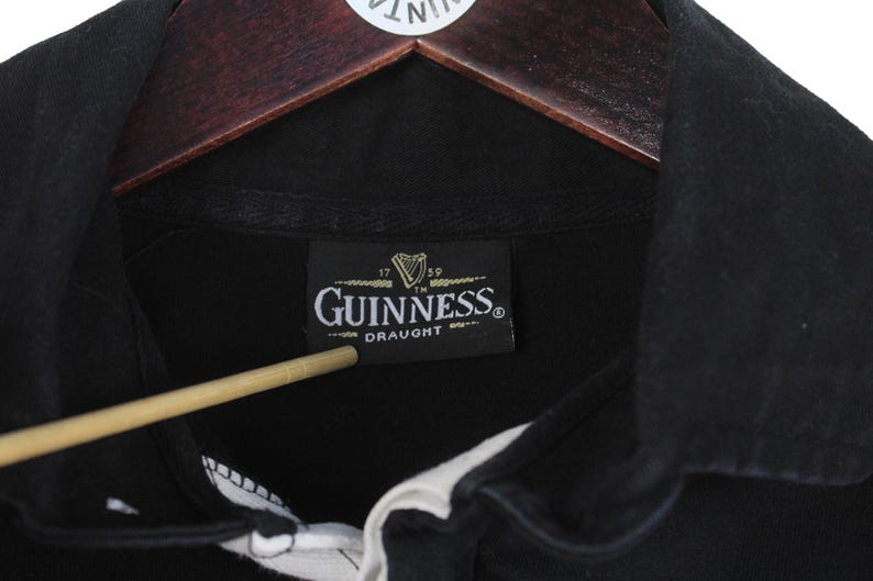 May include: Close-up of a black Guinness Draught shirt collar. The shirt has a white collar and a black label with the Guinness logo and text. The shirt is hanging on a wooden hanger.