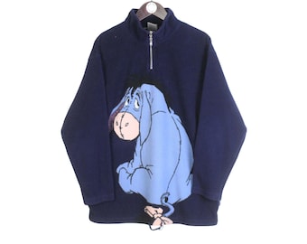 May include: Navy blue fleece pullover with a half-zip collar. The front features a large graphic of Eeyore, the donkey character from Winnie the Pooh, in shades of blue and pink. The pullover is hanging on a wooden hanger.