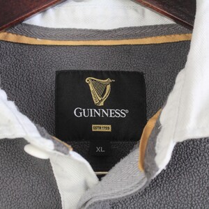 Puede incluir: Primer plano de un jersey de forro polar gris con cuello blanco. La etiqueta negra presenta el logotipo de Guinness y el texto "GUINNESS ESTD 1759". La camisa es talla XL.