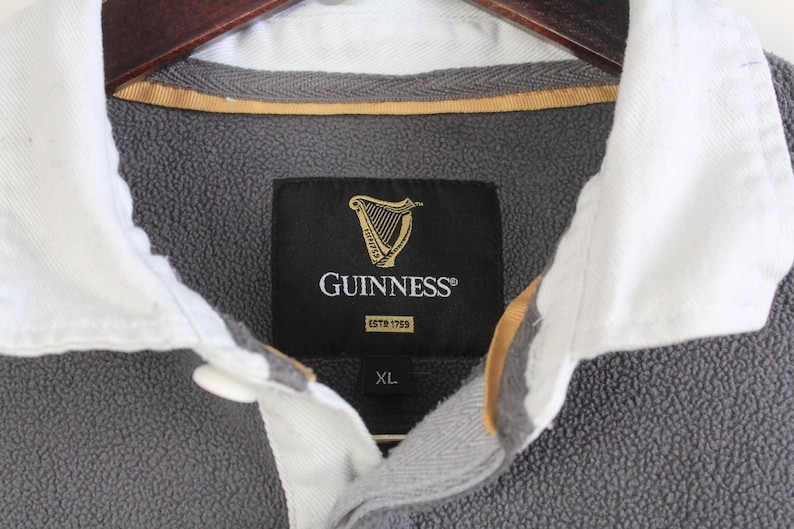 Puede incluir: Primer plano de un jersey de forro polar gris con cuello blanco. La etiqueta negra presenta el logotipo de Guinness y el texto "GUINNESS ESTD 1759". La camisa es talla XL.