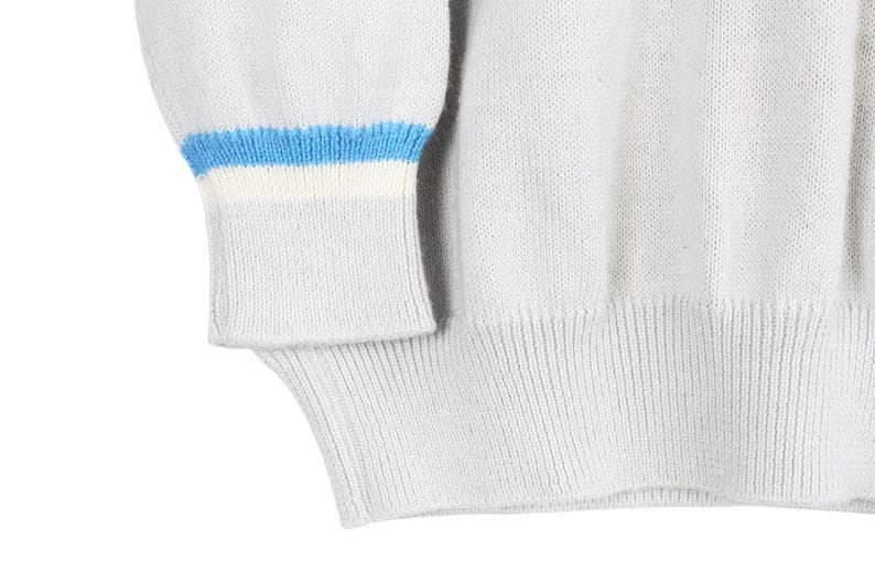 Puede incluir: Primer plano de un su&eacute;ter gris claro con dobladillo y pu&ntilde;os acanalados. El pu&ntilde;o presenta un dise&ntilde;o de rayas azules y blancas. El su&eacute;ter est&aacute; hecho de un material de punto suave.