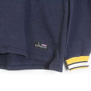 K&ouml;nnte beinhalten: Marineblaues Sweatshirt mit gelb-wei&szlig; gestreiften B&uuml;ndchen. Das Kangol-Logo ist auf dem Kleidungsst&uuml;ck sichtbar. Das Sweatshirt ist aus einem weichen Material gefertigt.