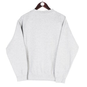 Puede incluir: Una sudadera gris claro con cuello redondo y mangas largas, colgada de una percha de madera. La sudadera tiene un dise&ntilde;o sencillo y cl&aacute;sico, adecuado para ropa informal. La tela parece ser un material suave y c&oacute;modo.
